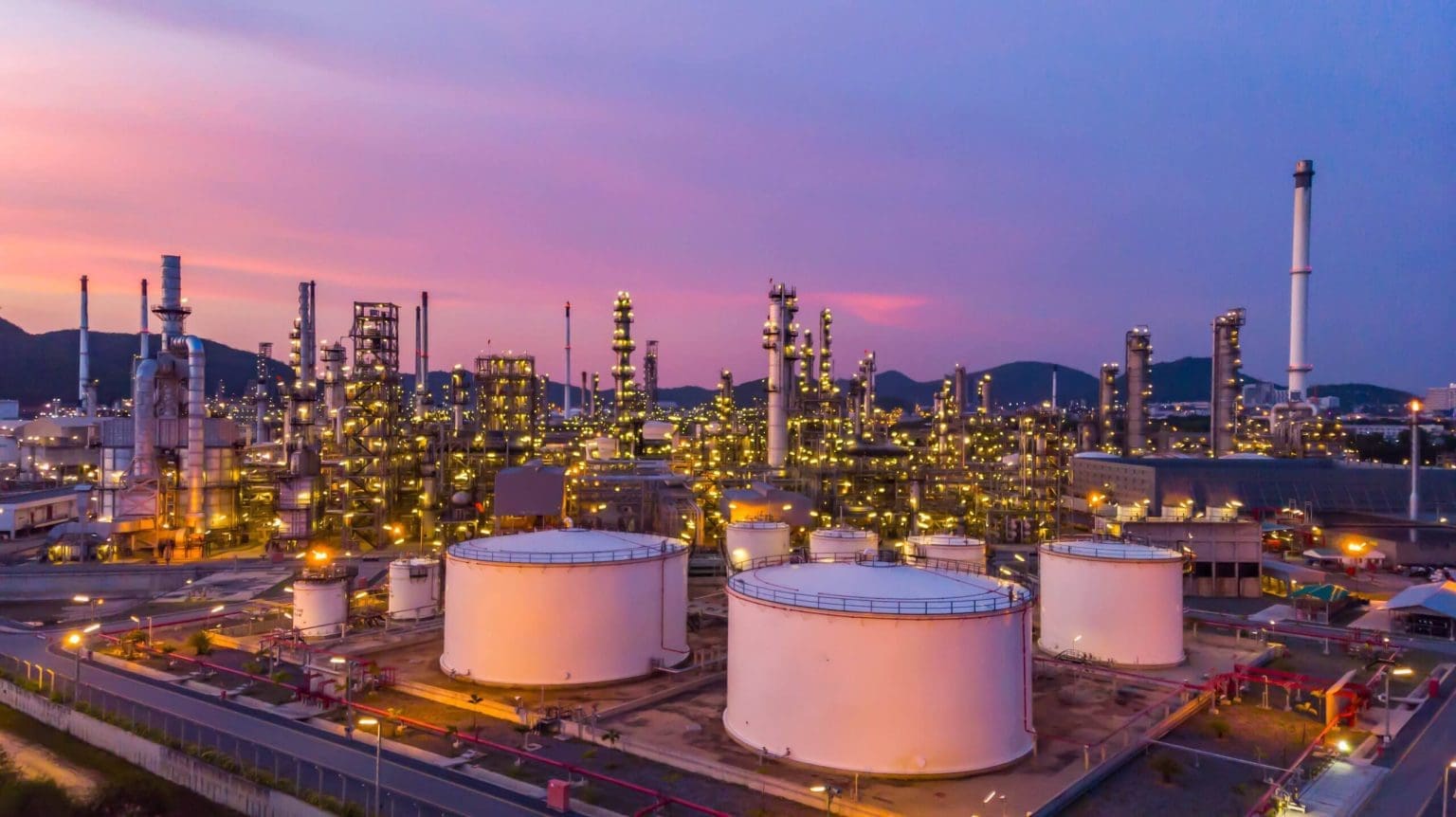 Aerial view of a state-of-the-art oil refinery at dusk, featuring illuminated storage tanks, distillation towers, and interconnected pipelines against a vibrant sunset sky.