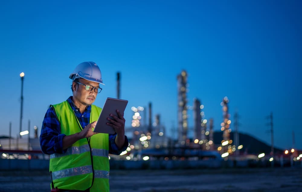 An industrial worker in safety gear using a tablet at a refinery site during dusk, representing the use of technology in monitoring and managing industrial operations.