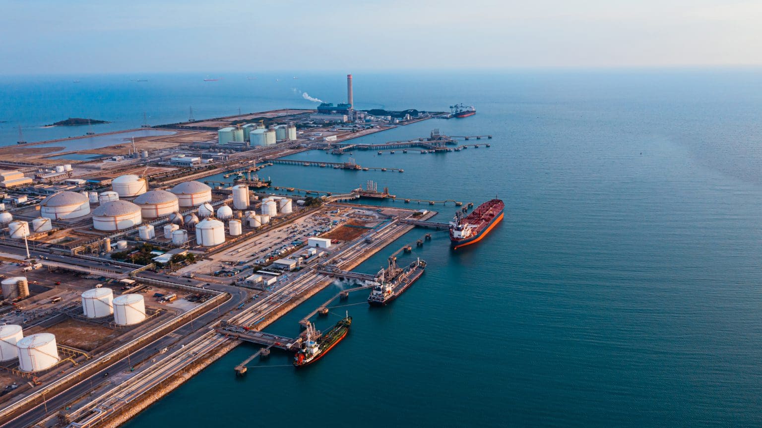 Aerial view of an oil terminal with large storage tanks, docked tanker ships, and extensive infrastructure along the coastline.